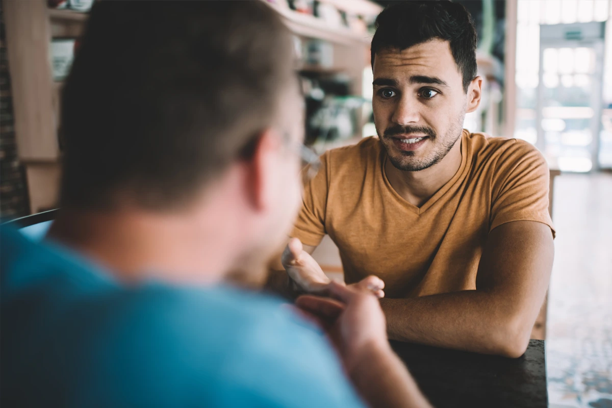 A group therapy session with three men seated in a circle, engaging in a supportive conversation in a bright room.