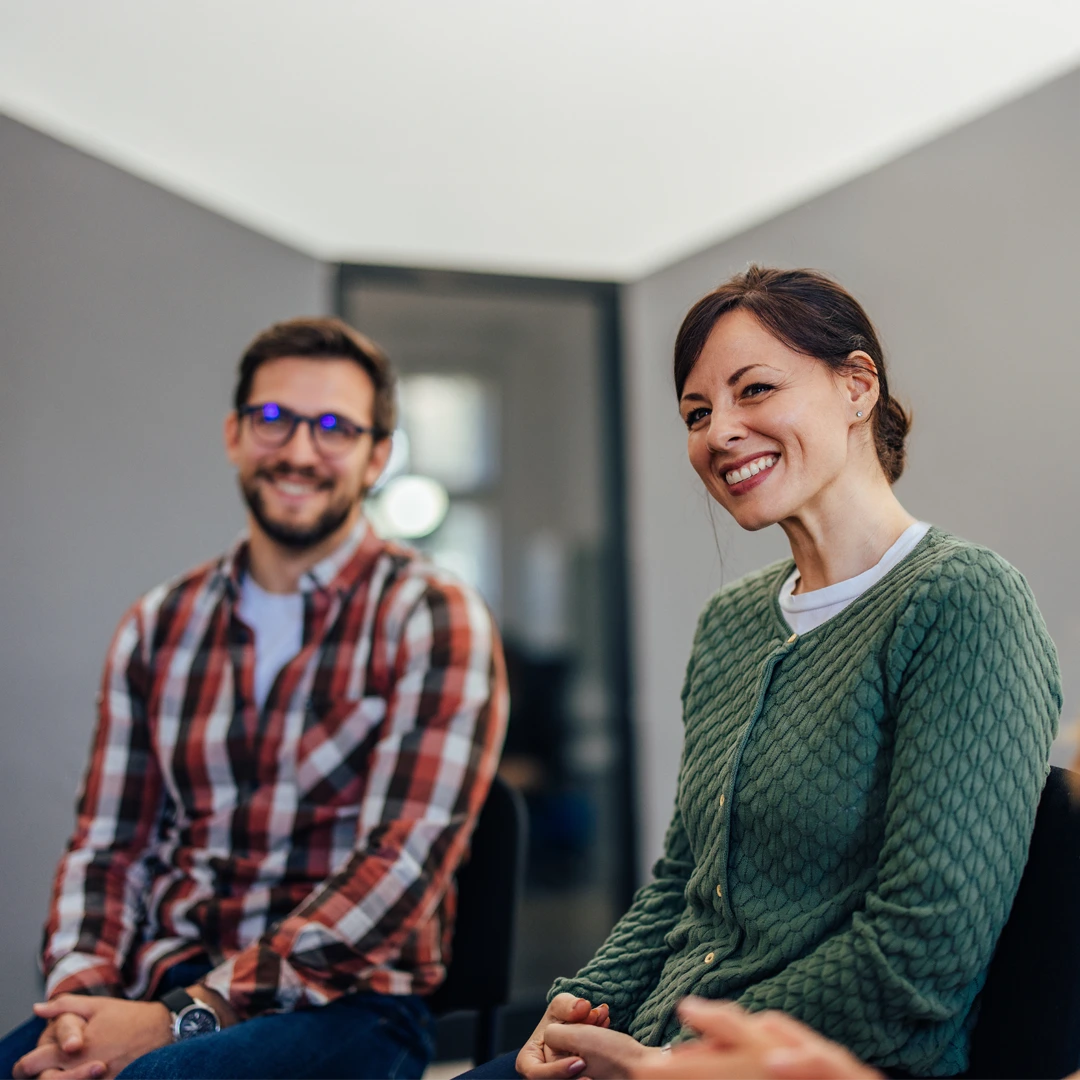 Smiling woman and man seated in a softly lit room, appearing engaged in a supportive conversation during a therapy or group session.