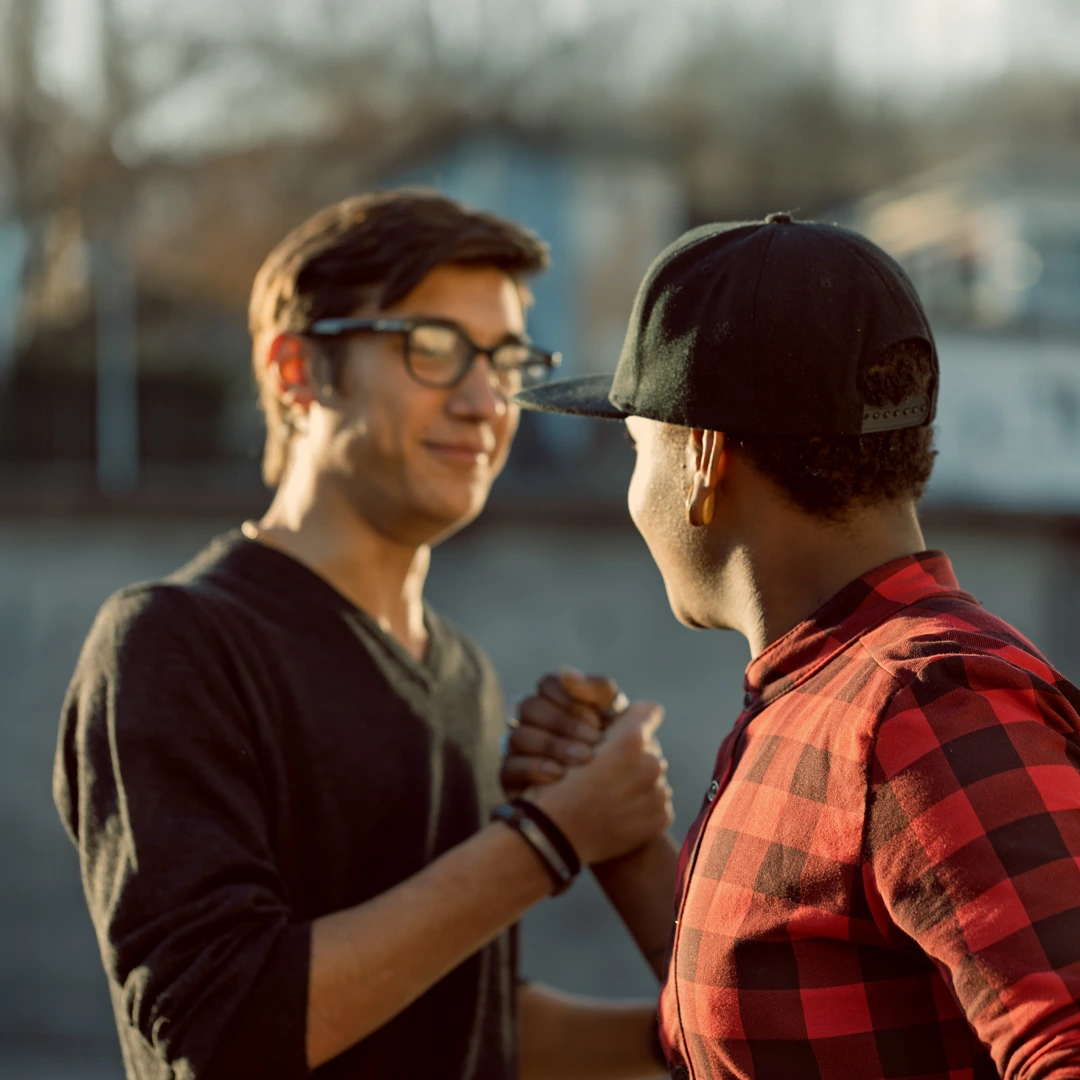 Two young men greeting each other with a friendly handshake and smile outdoors, symbolizing connection, trust, and mutual support.