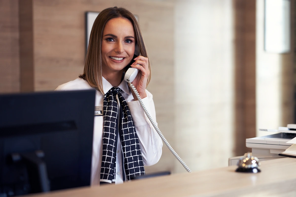 Woman receptionist smiling while speaking on a landline phone at a welcoming front desk.