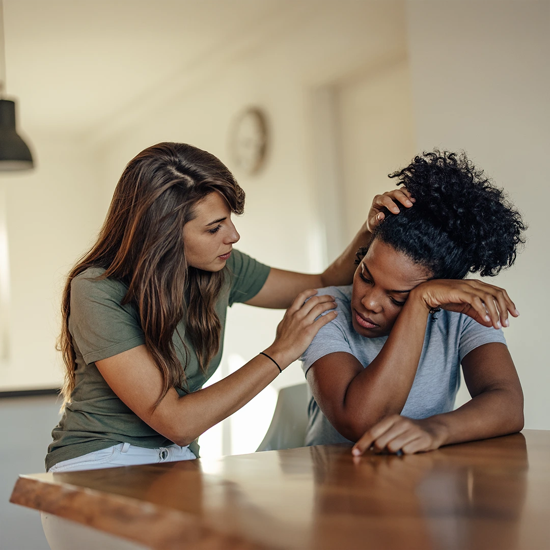 A supportive young woman gently comforts another woman who appears sad and overwhelmed at a table, symbolizing compassion and emotional support.