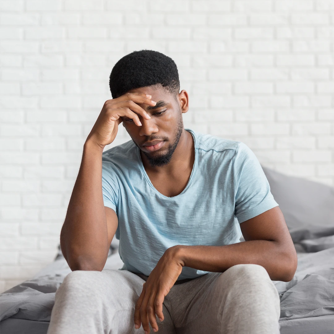 Man sitting on bed with head resting on hand, appearing overwhelmed and distressed, representing the emotional burden of heroin addiction.