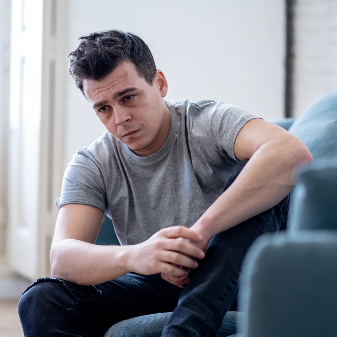A young man in a gray t-shirt sits on a couch looking distressed and deep in thought, symbolizing the emotional struggle of addiction.