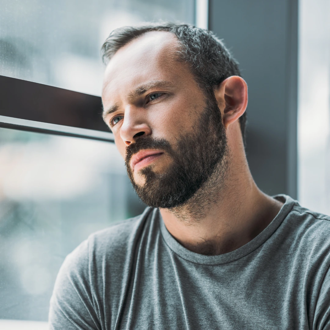 Bearded man in a gray shirt leaning against a window, appearing lost in thought, representing the challenges of heroin addiction.