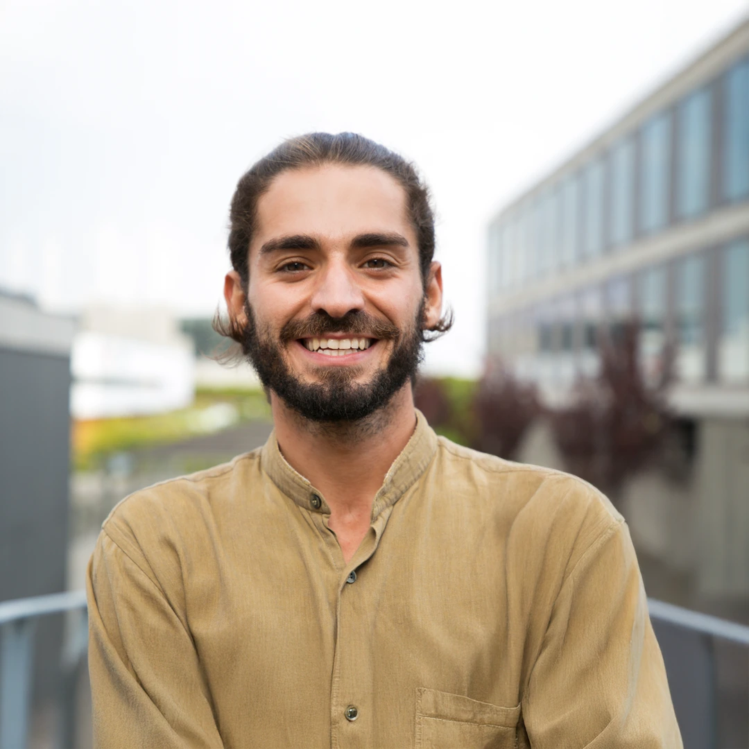 Smiling young man with beard and tied-back hair standing outdoors in a modern urban setting.