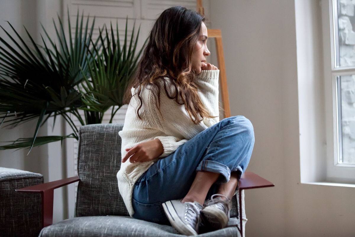 A-Closer-Look-at-Anxiety-Treatment-Mobile Woman sitting curled up on a chair, looking thoughtfully out a window, reflecting feelings of anxiety or deep contemplation.
