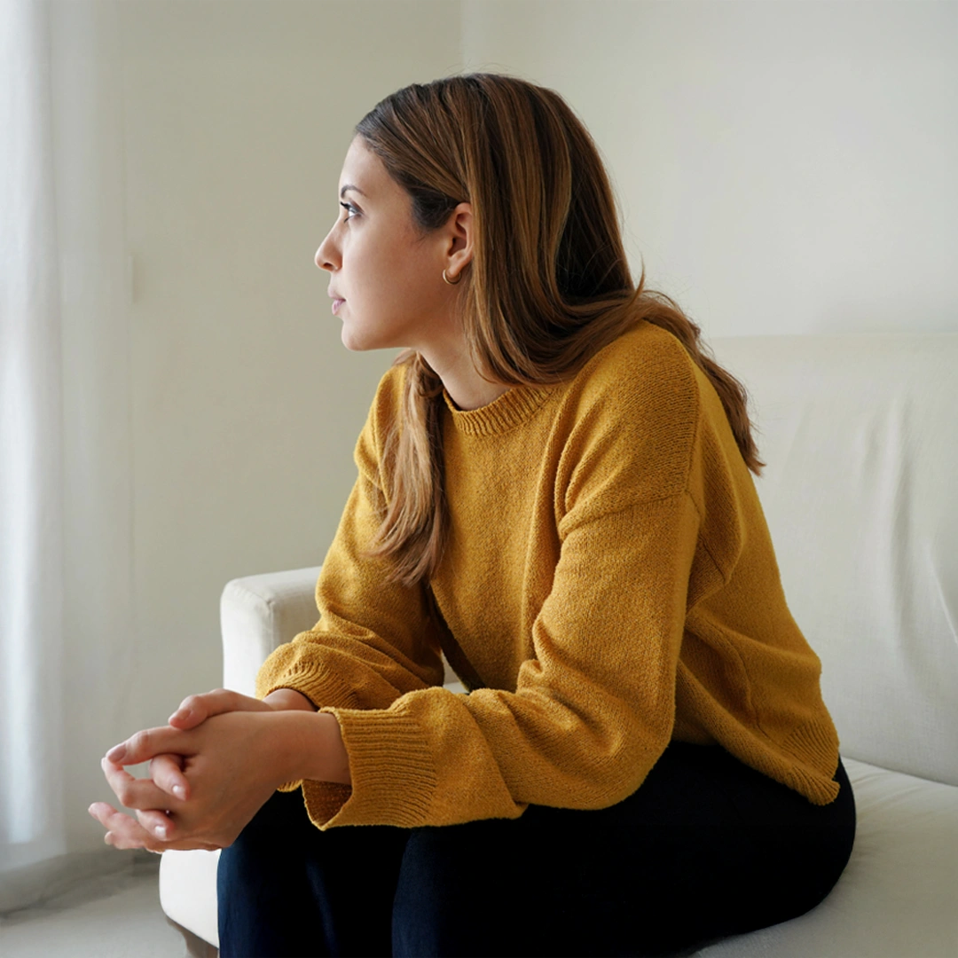 A-Closer-Look-at-Codependency-Treatment-Mobile Woman sitting thoughtfully on a couch in a bright room, looking out the window, representing self-reflection and emotional awareness during recovery from codependency.