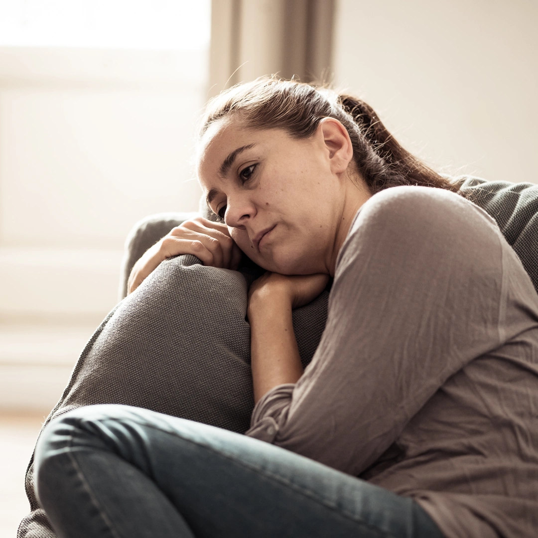 Woman sitting alone on a couch, resting her head on a pillow with a sad, distant expression.