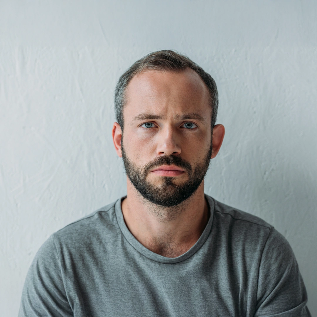 Serious young man with a beard wearing a gray t-shirt, looking directly ahead with a somber expression, symbolizing the emotional impact of meth addiction.