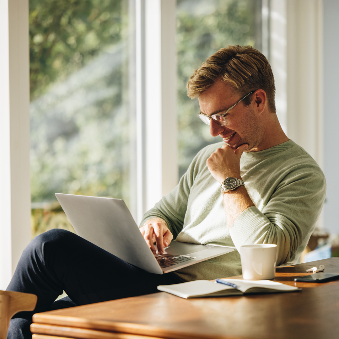 Comprehensive-Anxiety-Treatment-in-Arizona-Mobile Man sitting by a bright window working on a laptop, smiling thoughtfully with a coffee mug and notebook nearby.