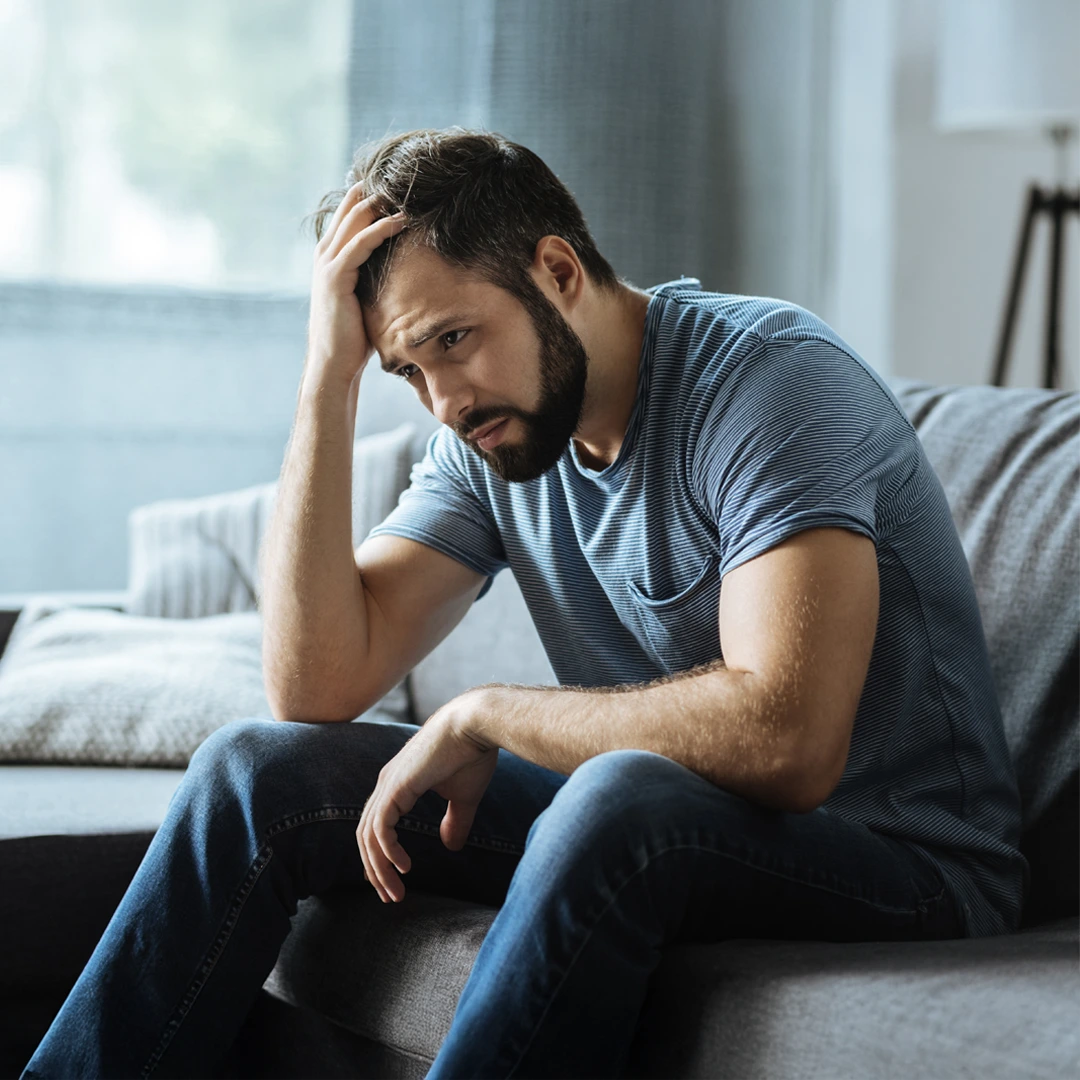 Comprehensive-Bipolar-Disorder-Treatment-Mobile Man sitting on couch with hand on head, appearing deep in thought or distressed, representing emotional challenges of bipolar disorder.