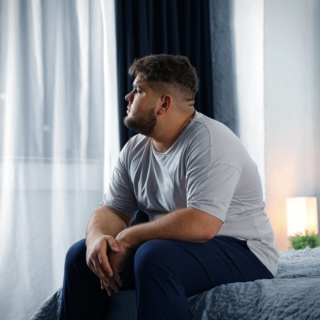 Comprehensive-Codependency-Treatment-in-Arizona-Mobile Man sitting on the edge of a bed looking out the window, appearing thoughtful and reflective, symbolizing the emotional challenges and isolation often experienced in codependent relationships.