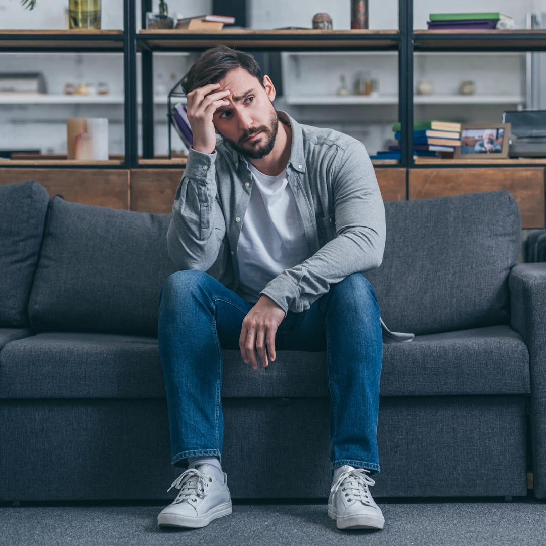 Man sitting on a couch with a thoughtful, distressed expression, resting his head on his hand.