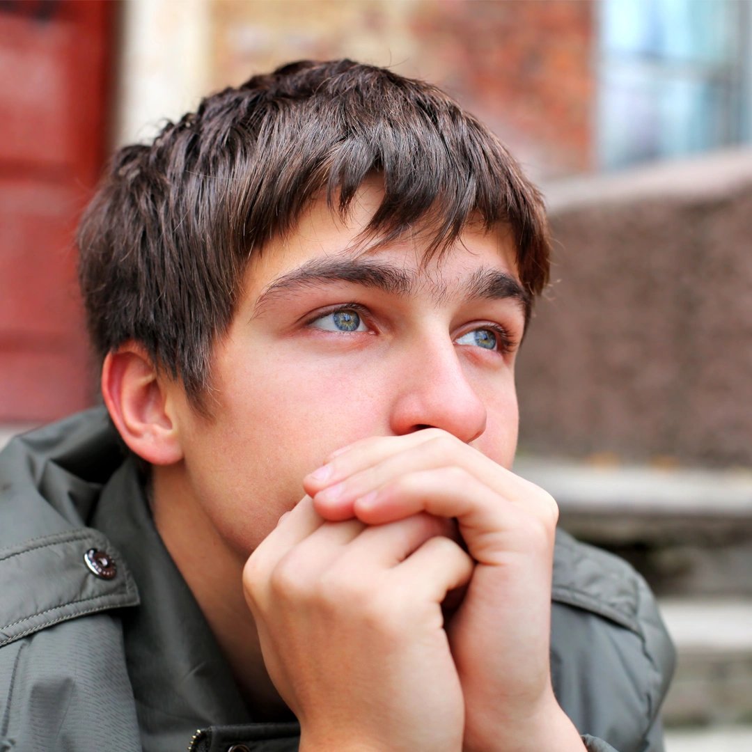 A young man sits outdoors with his hands clasped near his face, gazing upward with a thoughtful, emotional expression that conveys sadness and reflection.