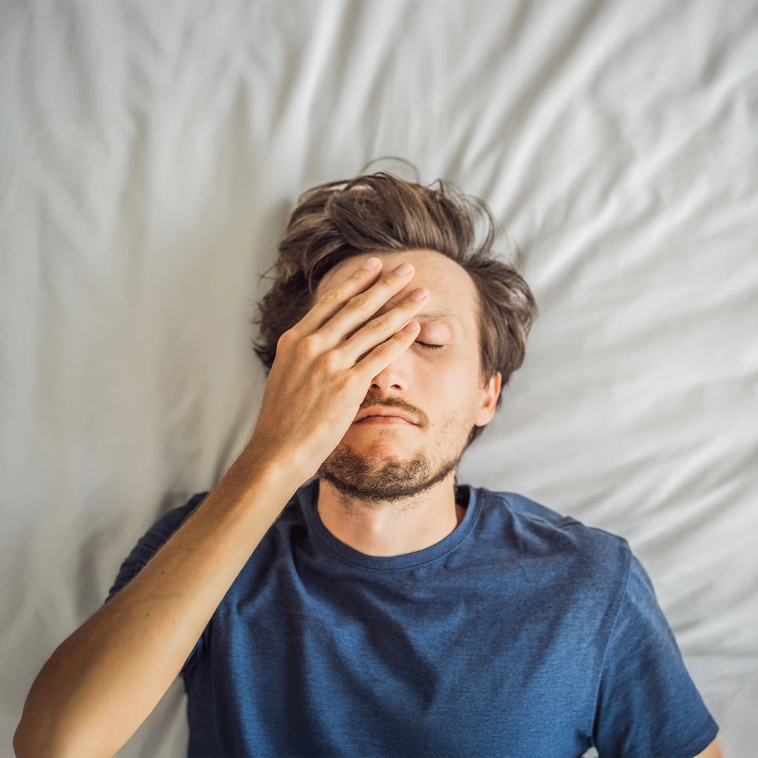Man lying on bed with his hand over his face, appearing distressed or exhausted, symbolizing the emotional toll of PTSD and the need for compassionate treatment.