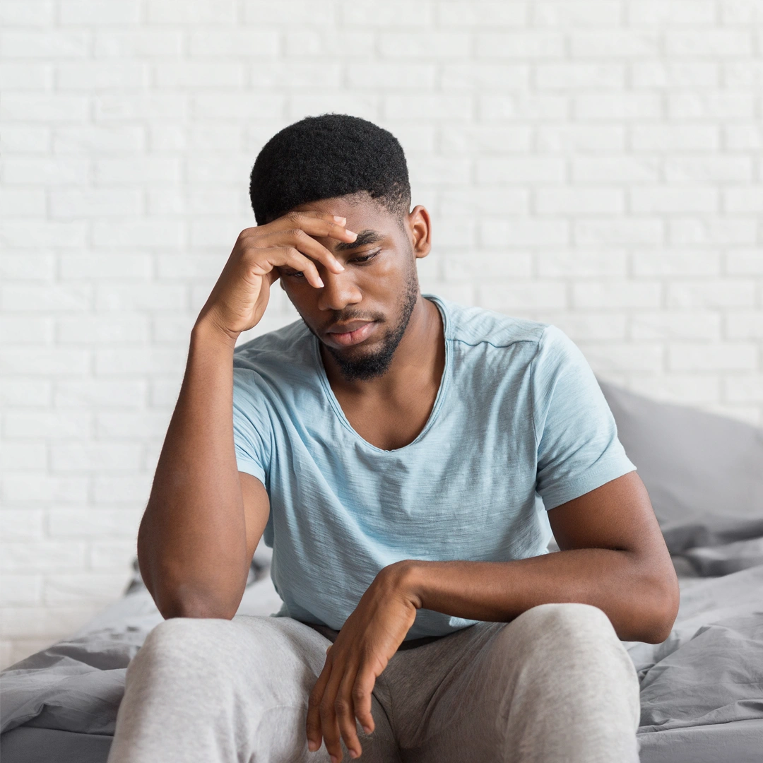 Young man sitting on a bed with head in hand, showing signs of stress and struggle, symbolizing the emotional toll of meth addiction.