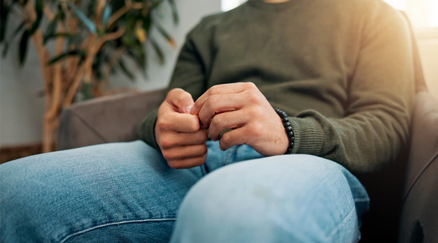 Close-up of a seated person wearing a green sweater and blue jeans, fidgeting with their hands while sitting on a couch, with a blurred indoor background and a plant visible.